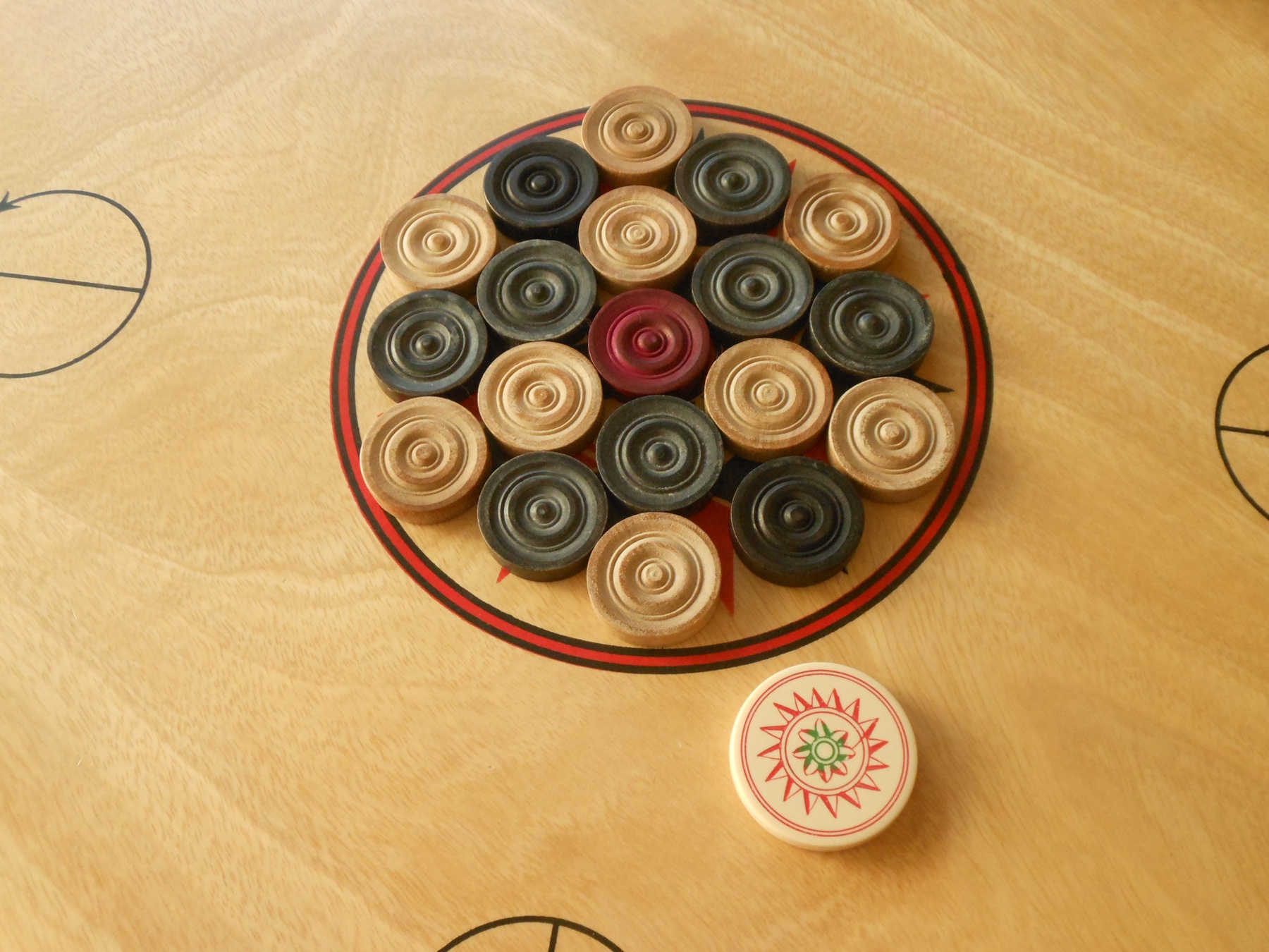 A carrom board set up for play, coins arranged around the centre with the red queen in the middle and the striker resting beside the ring